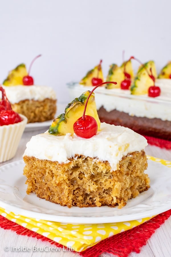 A white plate with a piece of pineapple sunshine cake topped with pineapple cream cheese frosting, a cherry, and pineapple slice on it