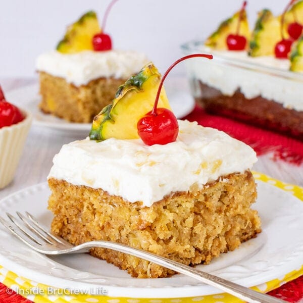 A white plate with a piece of pineapple cake topped with frosting, a cherry, and a pineapple wedge on it