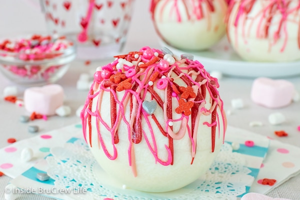 A white doily with a white hot chocolate bomb drizzled with red and pink chocolate and sprinkles on it