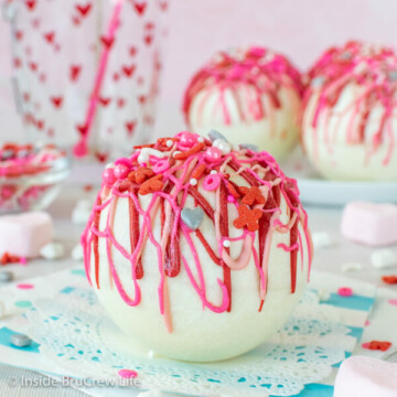 A strawberry white hot chocolate bomb with pink and red drizzles and sprinkles on a white doily