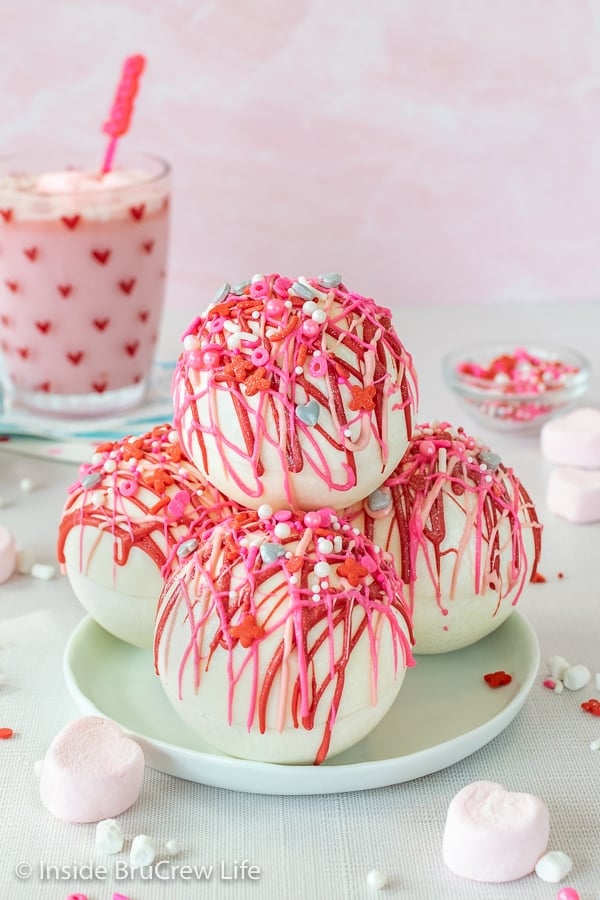 Four strawberry white hot chocolate bombs drizzled with pink and red chocolate and sprinkles stacked on a white plate