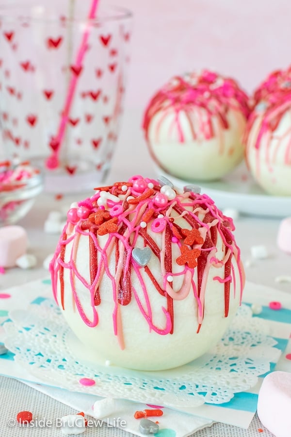 A white hot chocolate bomb filled with strawberry powder and drizzled with pink chocolate on a white doily