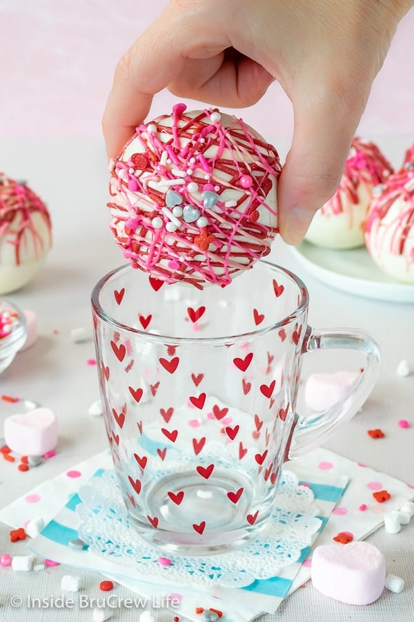 An empty clear mug with red hearts on a doily with a strawberry white hot chocolate bomb being held above it