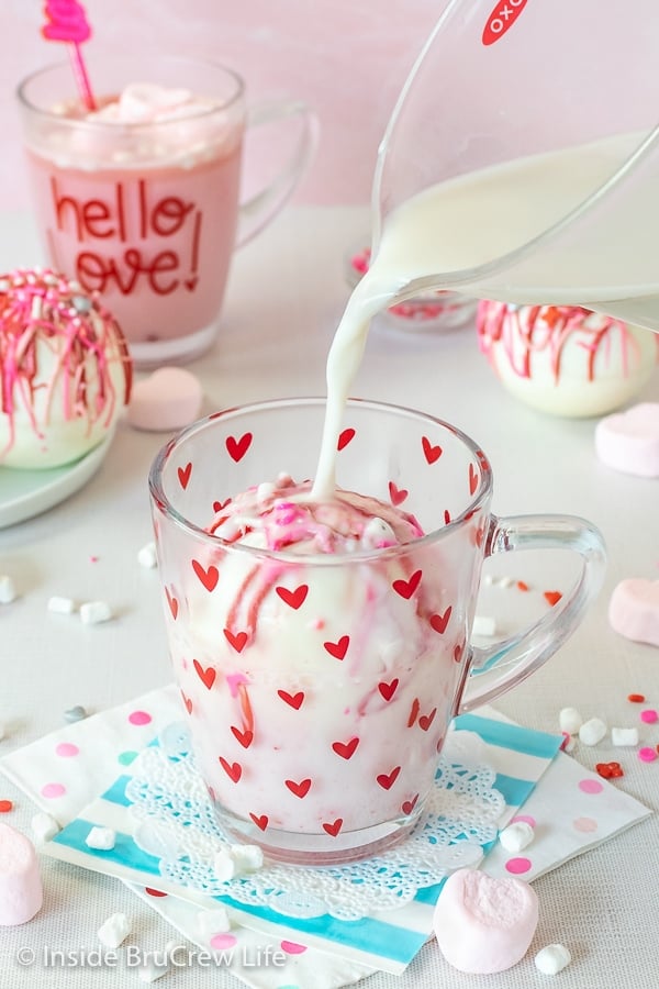 A clear mug with red hearts with a strawberry hot chocolate bomb in it and milk being poured over it