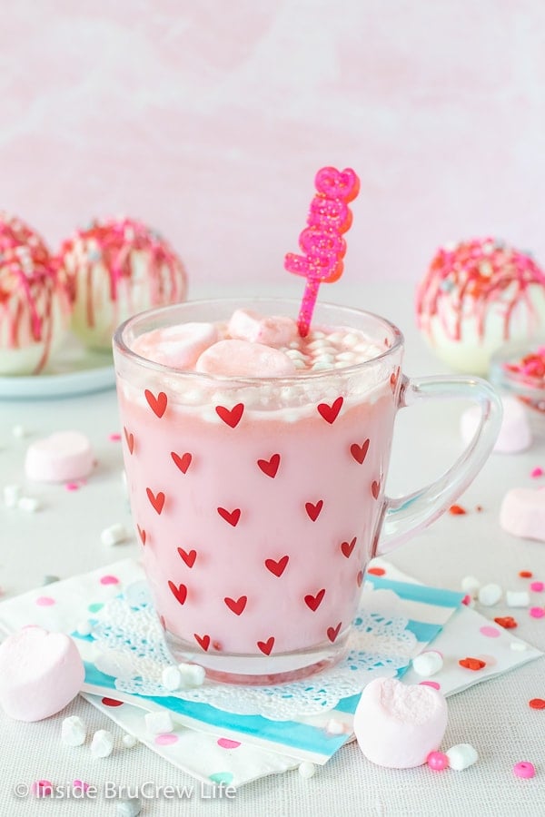 A clear mug with red hearts filled with strawberry hot chocolate with hot chocolate bombs behind it