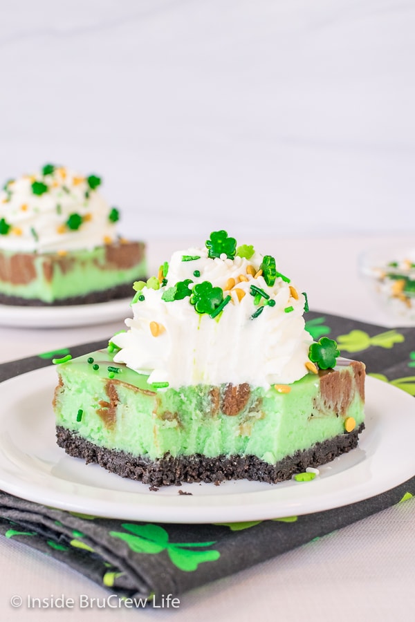 Two white plates with squares of mint cheesecake bars with chocolate swirls, whipped cream, and green sprinkles on them