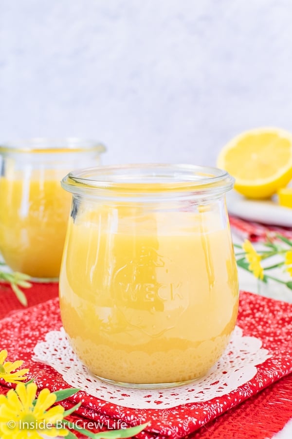 Two clear jars filled with lemon curd on a red towel with lemons behind them