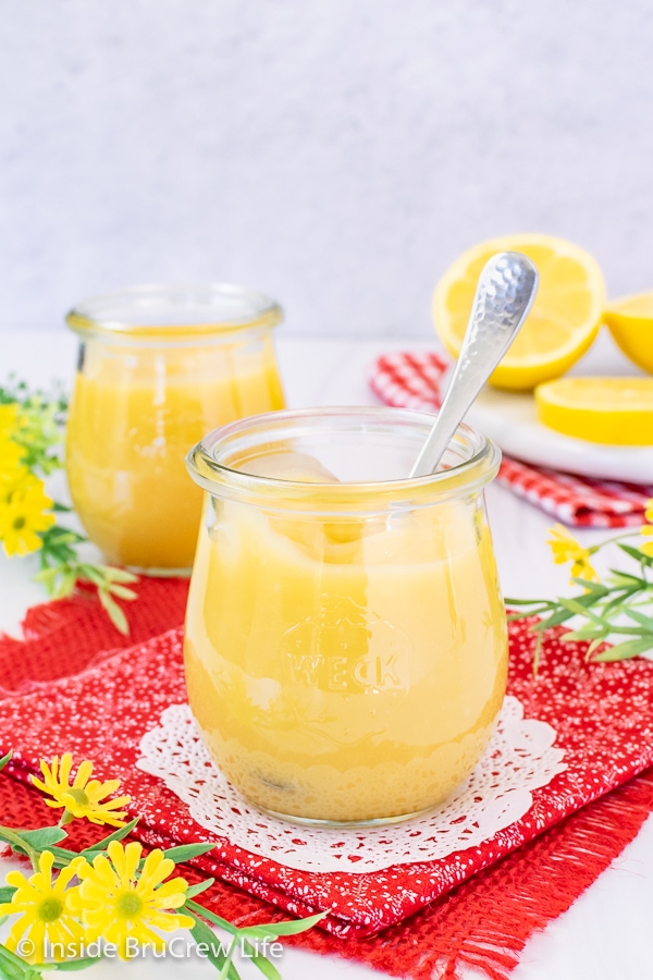 Two clear jars filled with lemon curd on a red towel with a spoon in one and lemons behind them