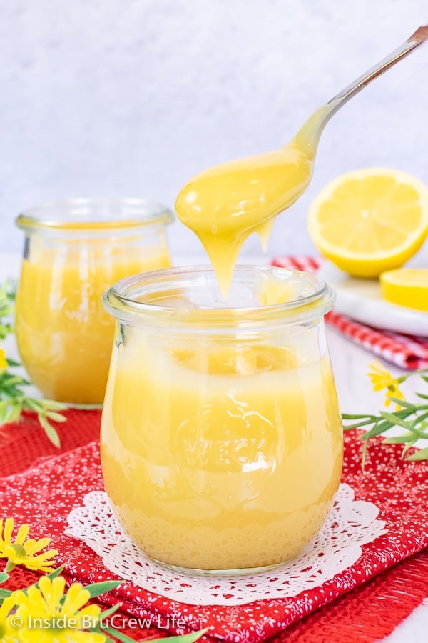 A clear jar on a red towel filled with easy lemon curd with a spoon lifting out a spoonful of sweet and tart goodness