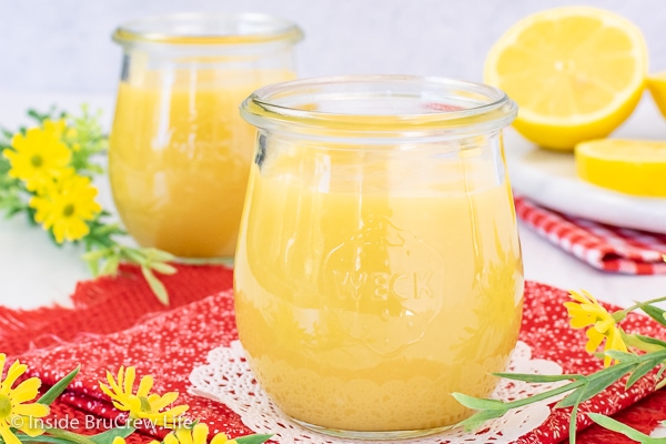Two clear jars filled with lemon curd on a red towel with lemons behind them