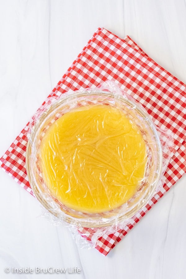 A bowl of easy lemon curd on a red and white towel with a piece of plastic wrap covering it