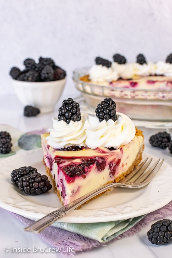 A white plate with a fork and a slice of cheesecake pie with blackberry swirls in it and whipped cream on top on it