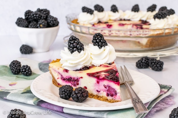 A white plate with a fork and a slice of swirled blackberry cheesecake pie with whipped cream on it