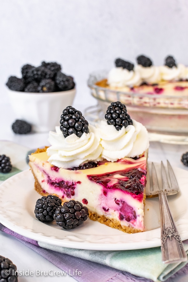 A white plate with a slice of blackberry cheesecake pie topped with whipped cream and fresh blackberries on it