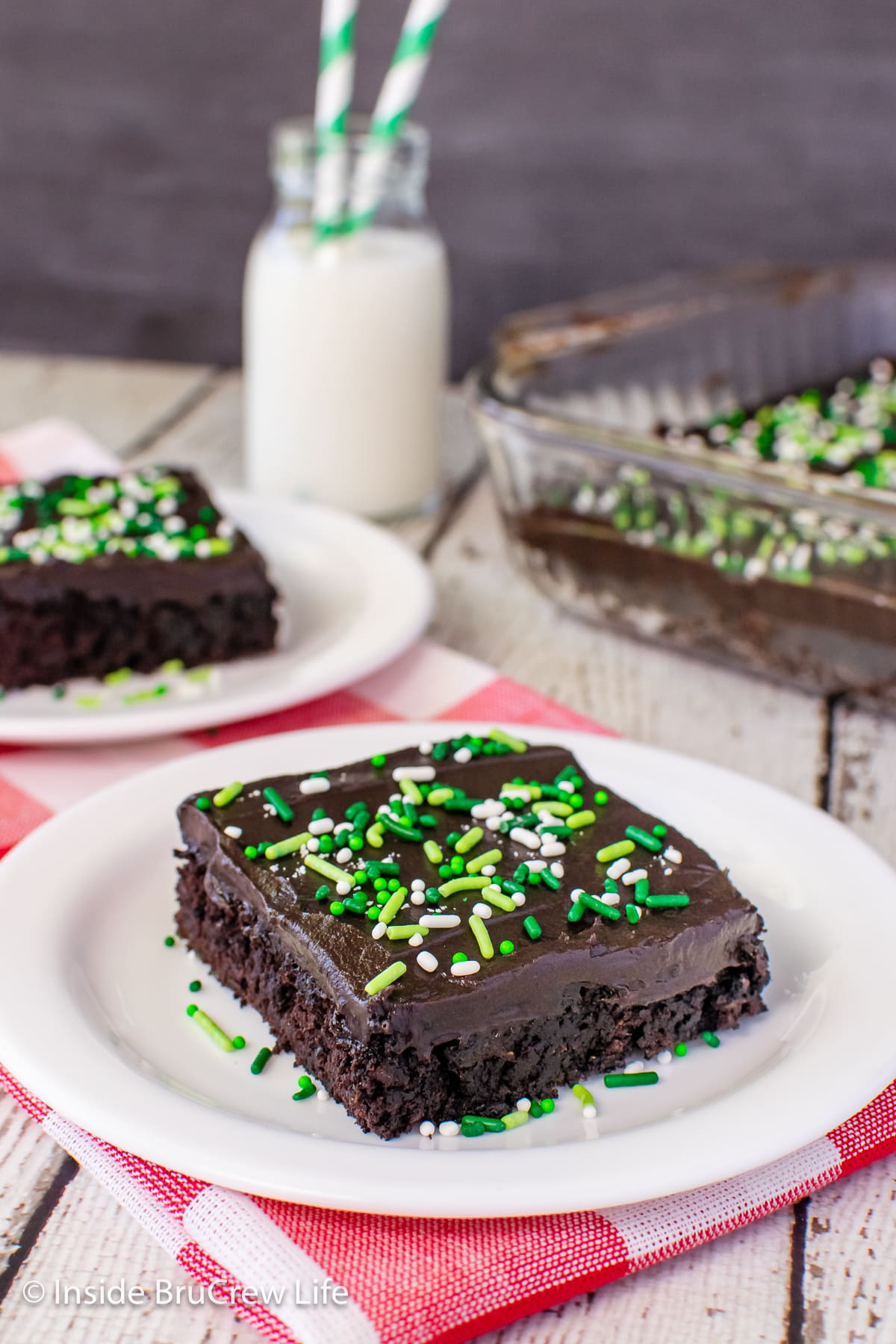 A white plate with a slice of frosted chocolate cake on it.