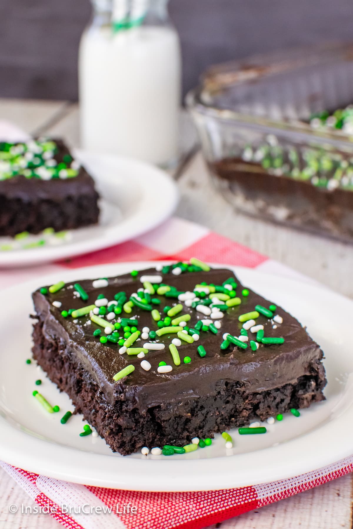 A square of chocolate cake with frosting and sprinkles on a white plate.