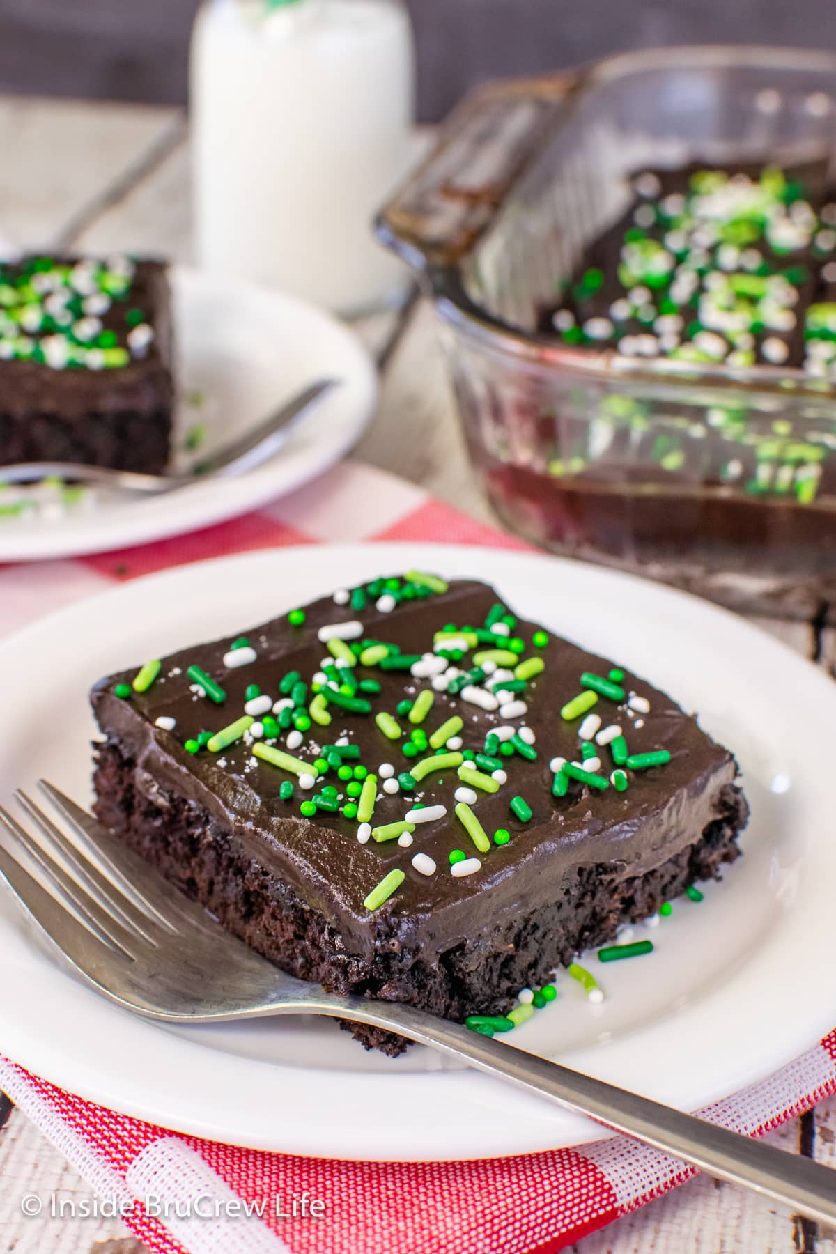 A slice of frosted chocolate cake with green sprinkles on a white plate.