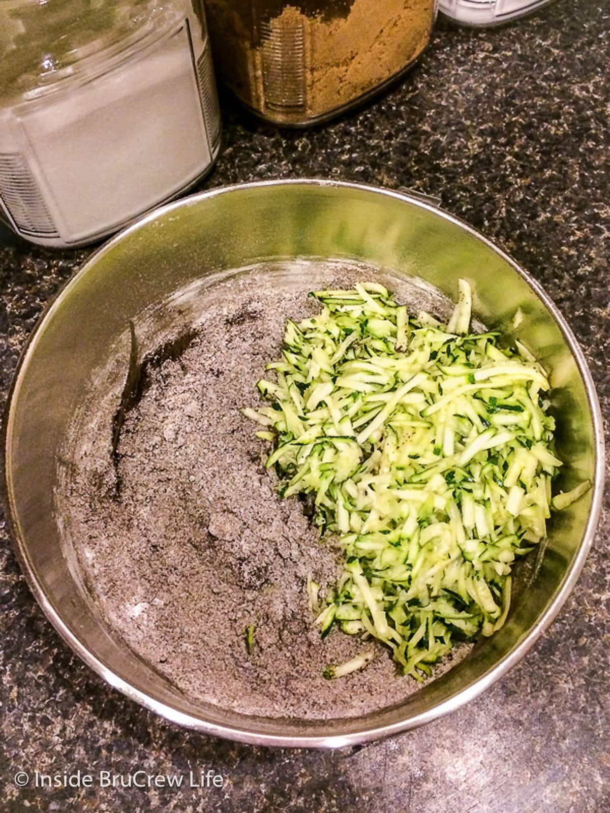 A metal bowl with ingredients needed to make a chocolate cake with shredded zucchini.