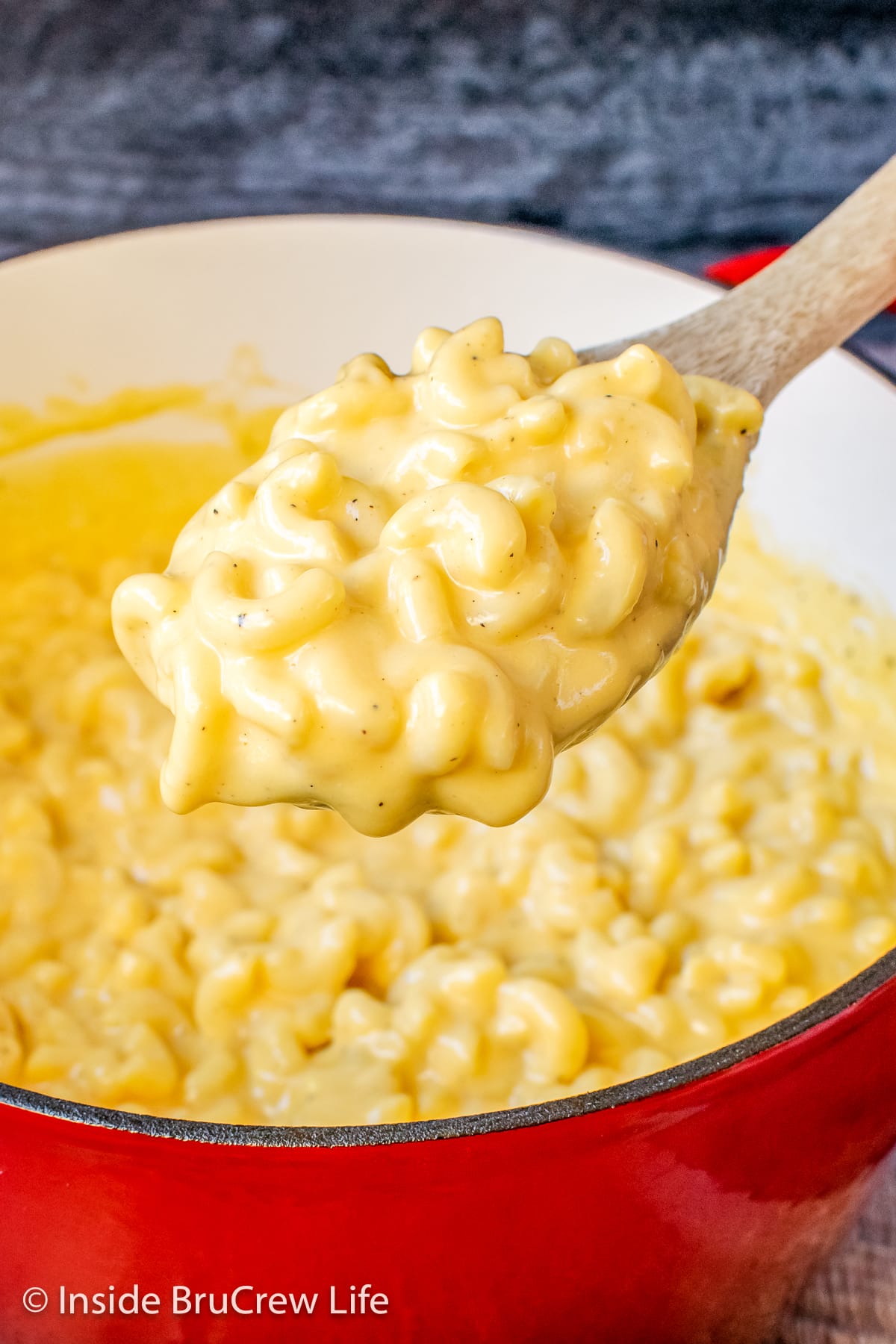 A wooden spoon lifting a scoop of creamy macaroni and cheese out of a red pot.