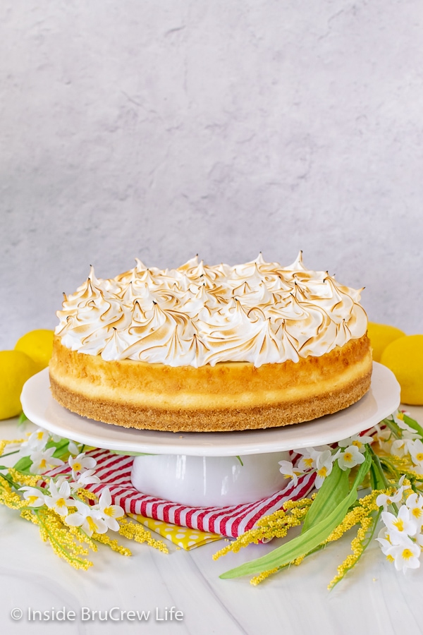 A white cake plate with a lemon cheesecake topped with toasted meringue topping on it.