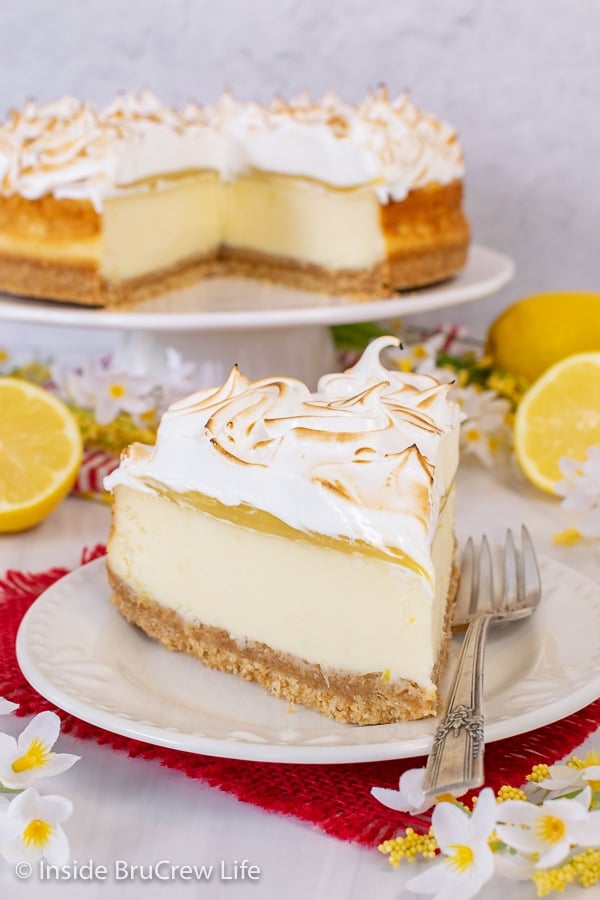 A white plate with a slice of lemon cheesecake topped with lemon curd and toasted meringue topping on it.