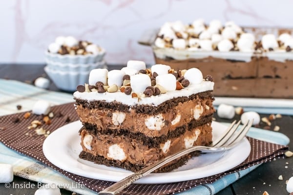 A white plate on a brown towel with a slice of rocky road icebox cake topped with marshmallows, nuts, and chocolate chips on it.