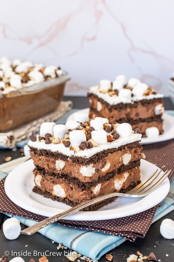 Two white plates with squares of chocolate icebox cake topped with marshmallows, nuts, and chocolate chips with a pan of dessert behind.