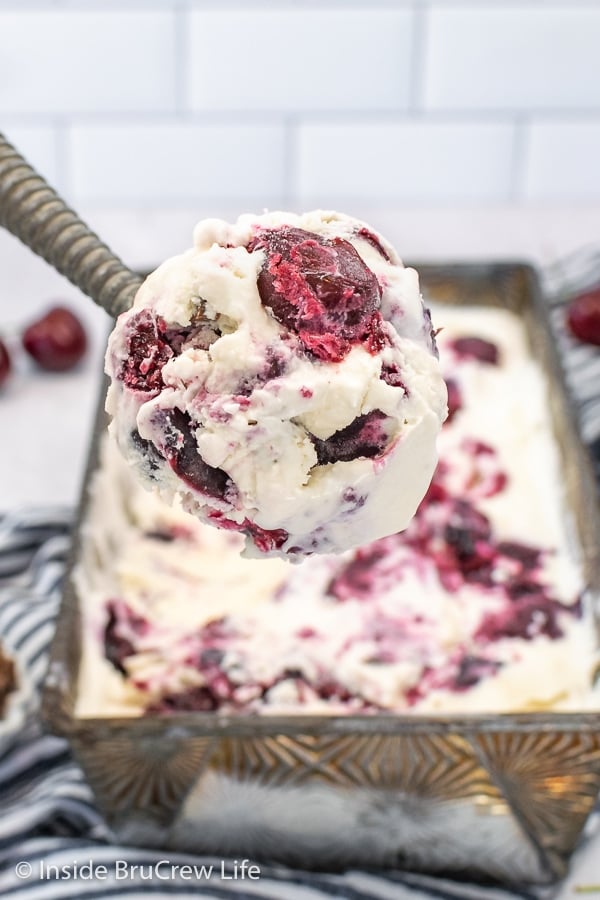 A pan of vanilla ice cream with cherries and a spoon lifting out a big scoop.
