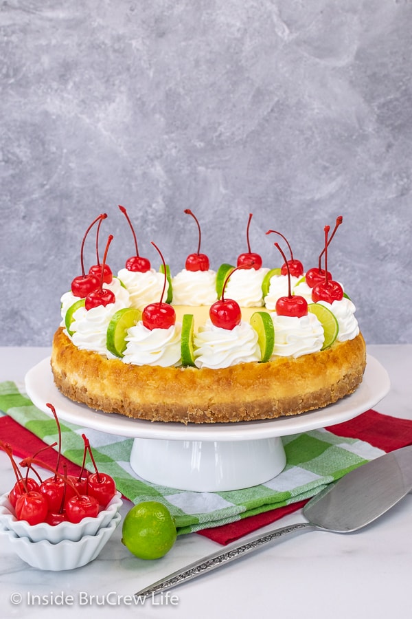 A white cake plate with a full sized key lime cheesecake topped with whipped cream, lime slices, and cherries.