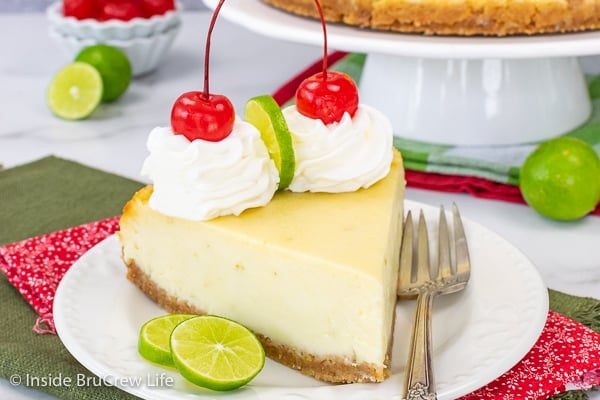 A white plate with a slice of key lime cheesecake topped with whipped cream, lime slices, and maraschino cherries.