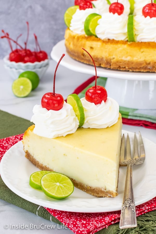 A white plate with a slice of key lime cheesecake with whipped cream and cherries on it.