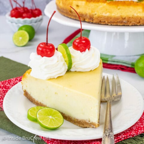 A white plate with a slice of key lime cheesecake with whipped cream and cherries on it.