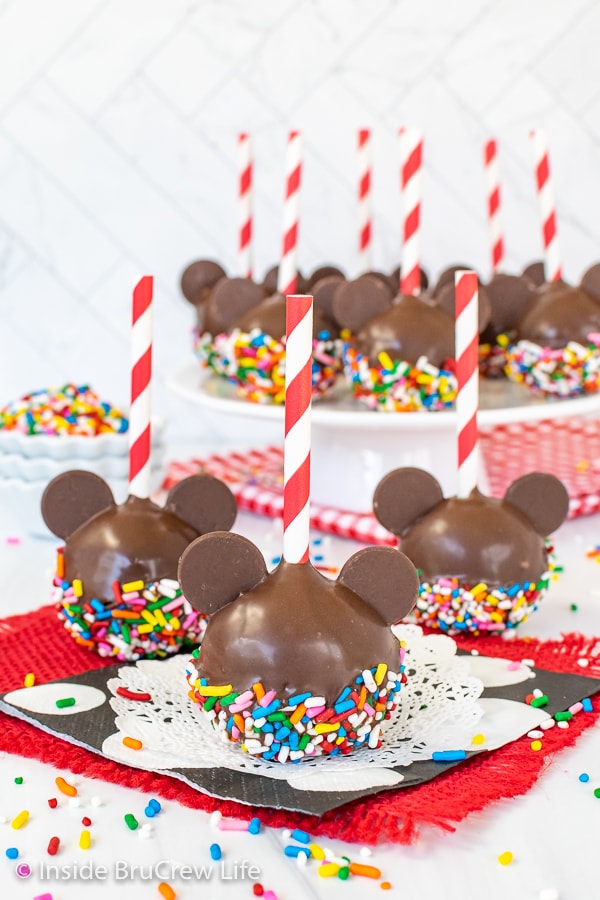 Three chocolate Mickey Mouse Cake Pops with sprinkles sitting on a black, white, and red napkin.