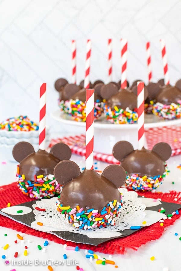 Three chocolate Mickey Mouse Cake Pops with sprinkles sitting on a black, white, and red napkin.