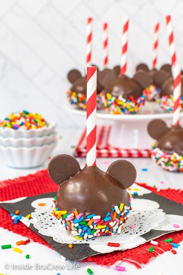 One Mickey Mouse Cake Pop with colorful sprinkles on a white doily and a cake plate with more chocolate cake pops behind it.