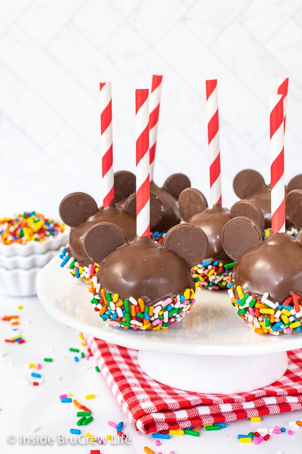 A white cake plate with seven Mickey cake pops with red and white straws on it.