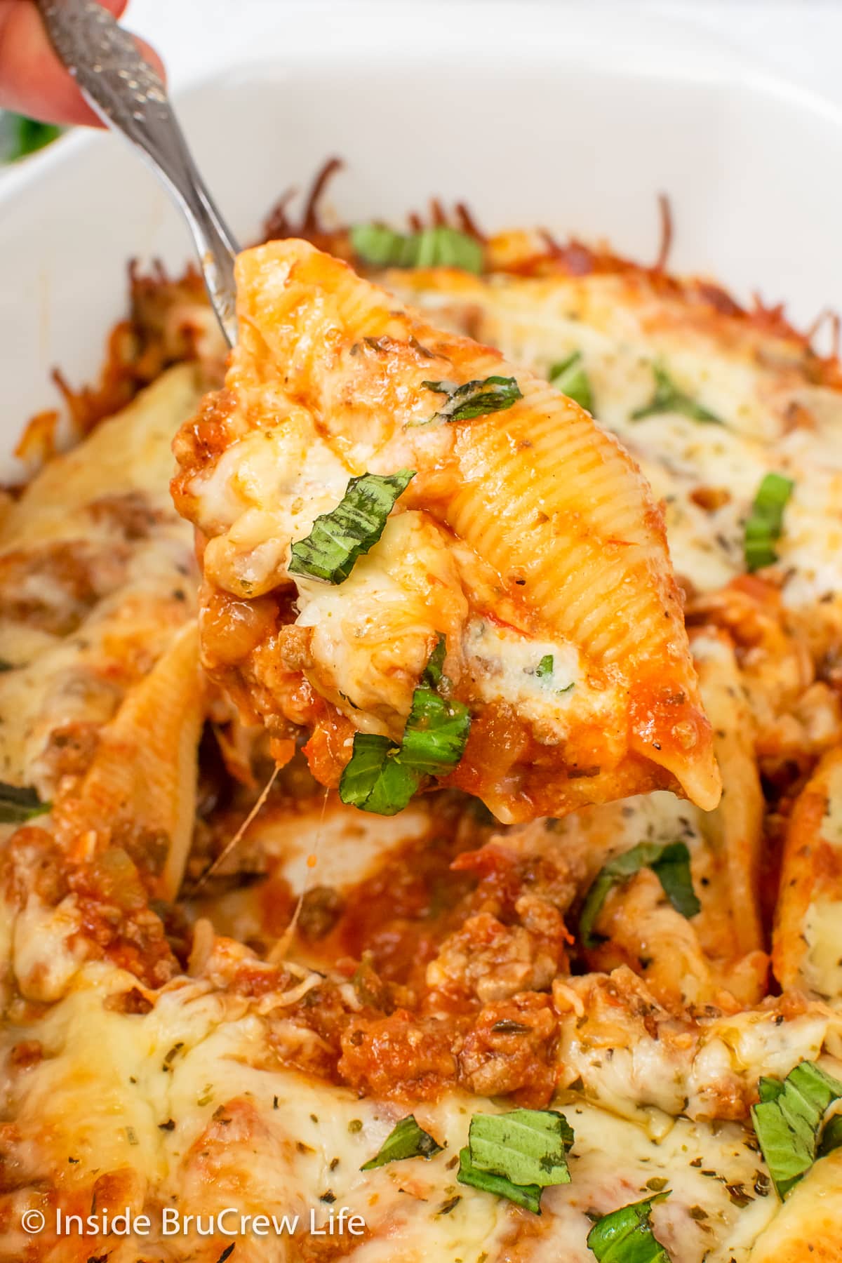 A spoon lifting a cheese and meat stuffed shell out of a casserole dish.