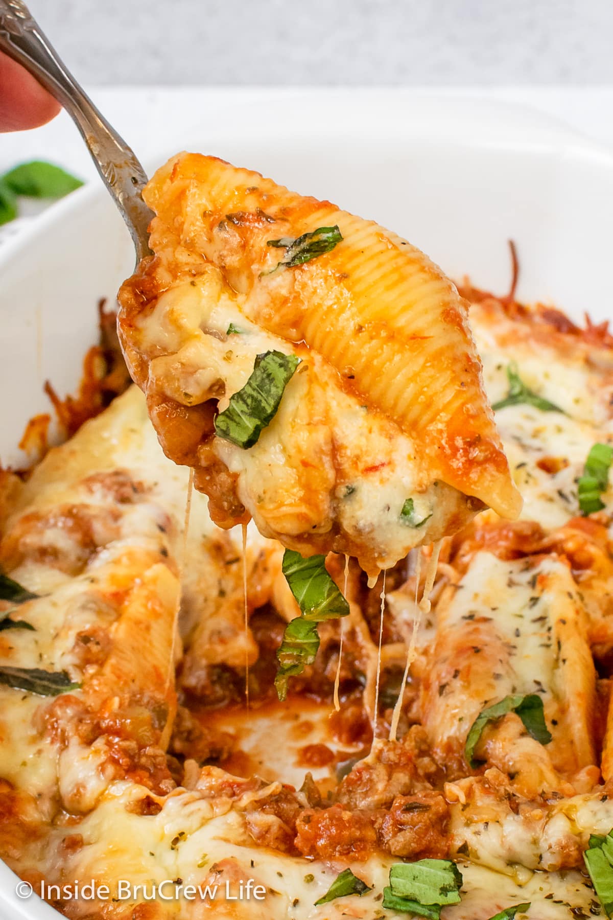 A spoon lifting a cheesy pasta shell out of a white casserole dish.