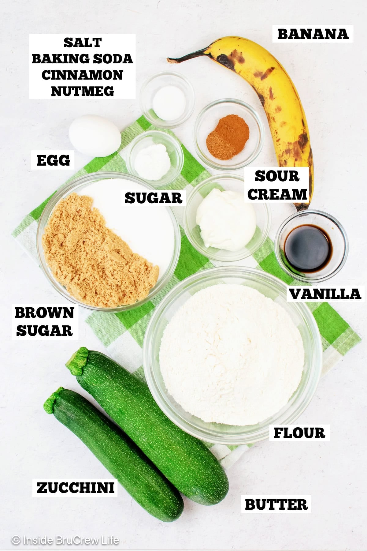 Ingredients needed to make a banana zucchini cake on a white board with labels.