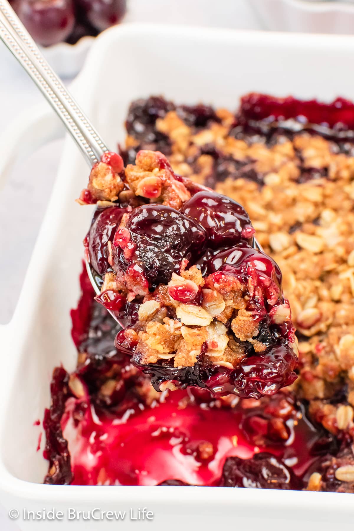 A white pan filled with cherry crisp and a spoon lifting some out.