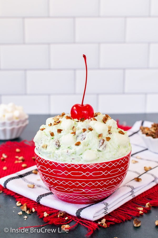 A red bowl filled with green pistachio fluff and topped with pecans and a maraschino cherry.