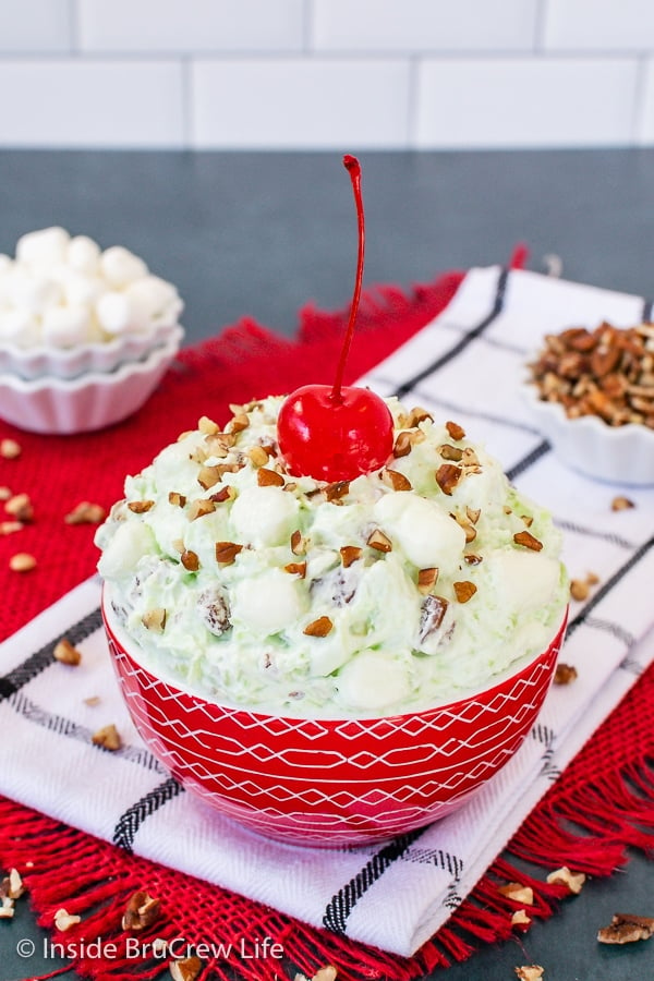 Green pistachio fluff salad in a red bowl topped with pecan chips and a red cherry with a stem.