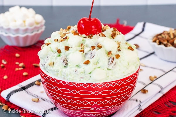 A red bowl on a white towel filled with pistachio salad and topped with pecans and a cherry.
