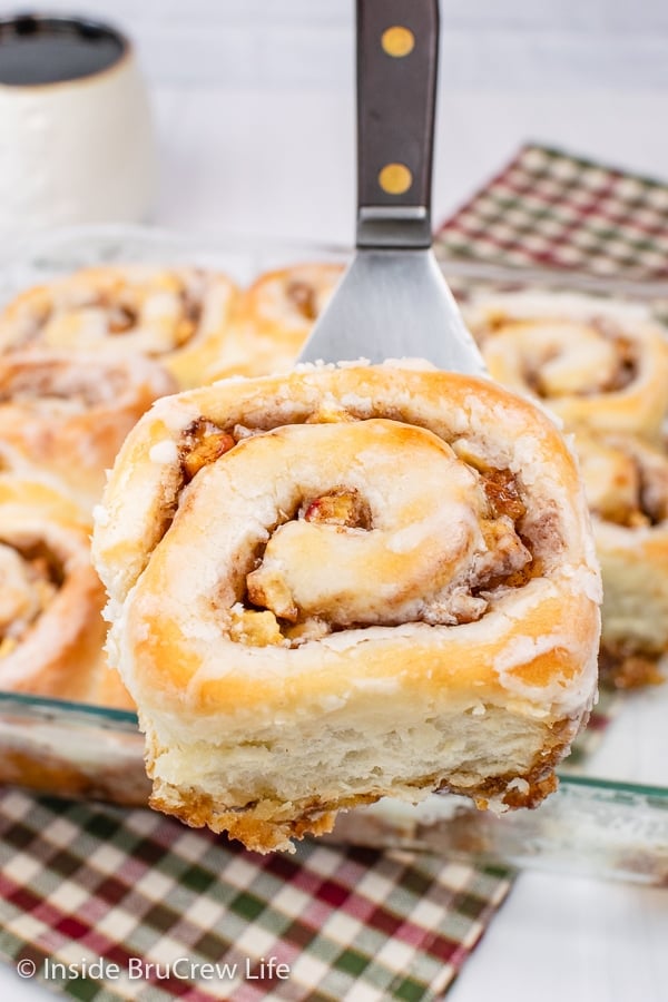 A metal spatula lifting an apple cinnamon roll out of a pan.