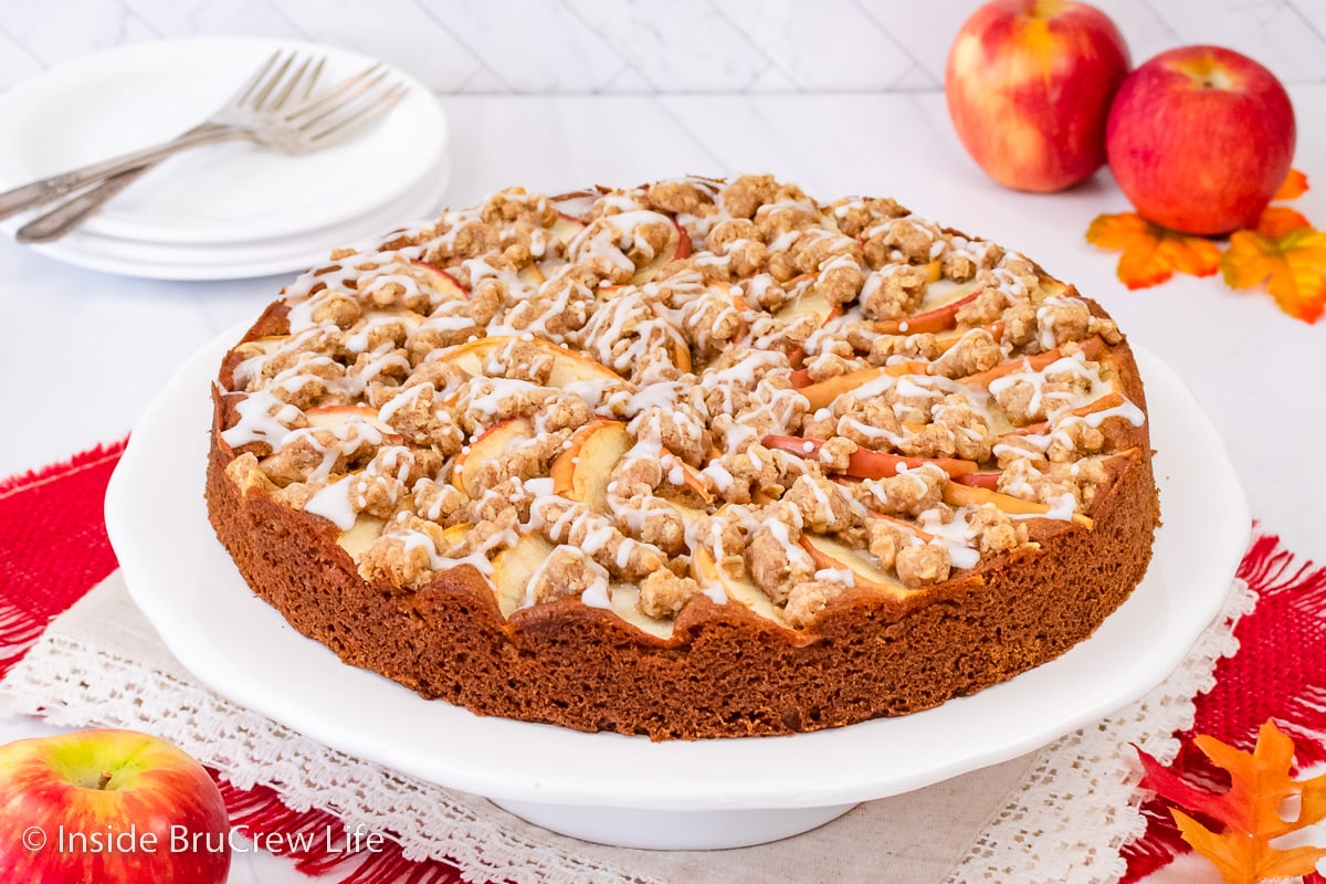A white cake plate with a full apple cake on it.