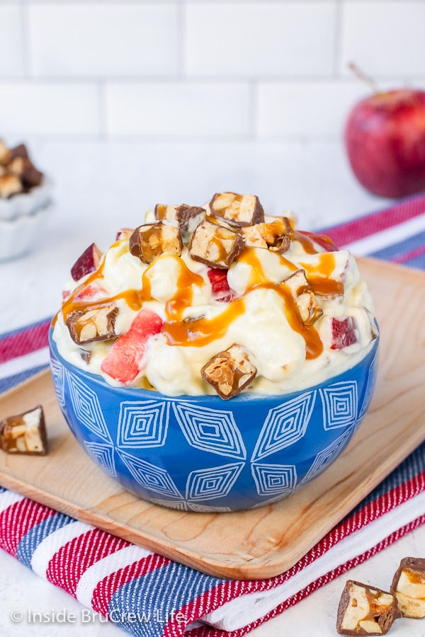 A blue bowl on a wooden board filled with apple snickers salad and topped with more candy bars and more caramel.