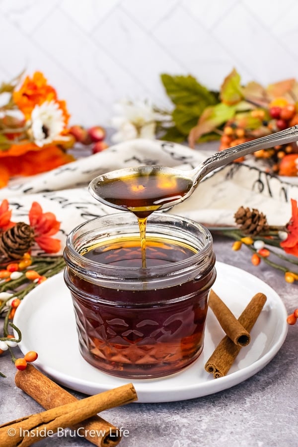 A clear jar on a white plate filled with cinnamon syrup with a spoon drizzling more syrup into it.