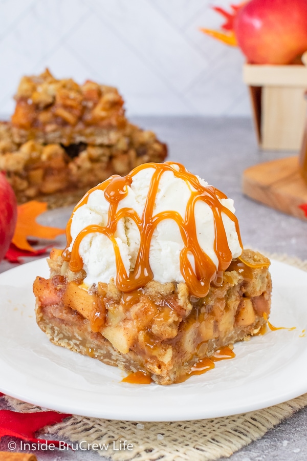 A white plate with a square of caramel apple pie bars topped with vanilla ice cream and caramel drizzles.