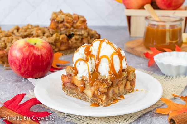 A white plate with a piece of caramel apple pie bars topped with vanilla ice cream and caramel drizzles.