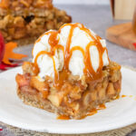 A white plate with a square of apple pie bars topped with vanilla ice cream and caramel.